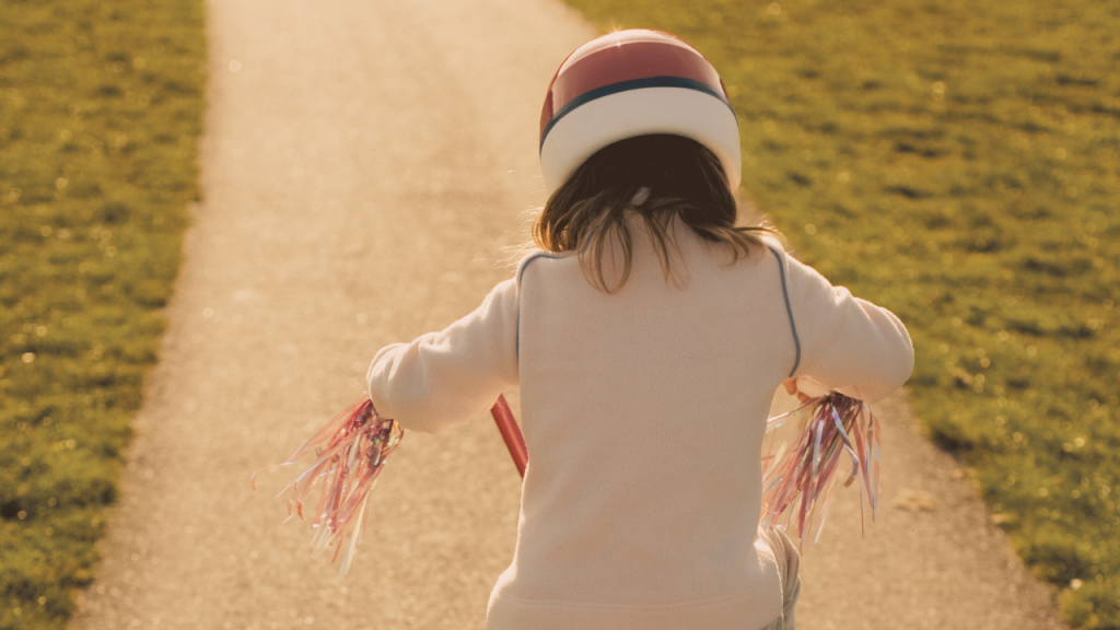 A young child rides a small bicycle down a quiet suburban street, tassels flying from the handlebars. The image symbolizes childhood independence, emotional growth, and a mother’s journey of raising kids while reparenting herself.